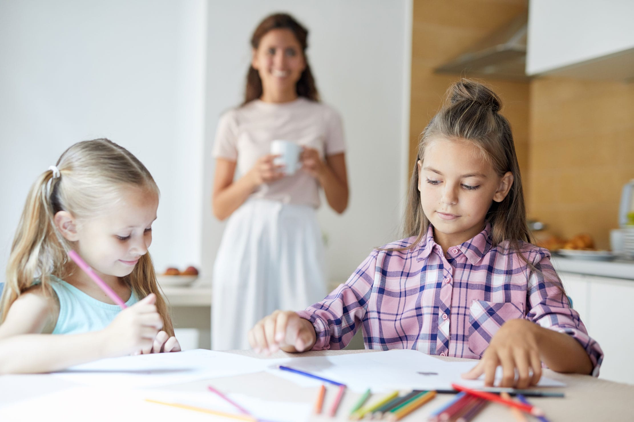 Youthful girls with crayons drawing pictures while sitting by table in the kitchen with their mom on background