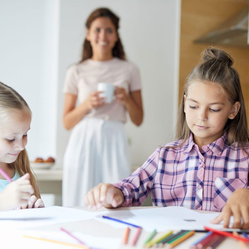 Youthful girls with crayons drawing pictures while sitting by table in the kitchen with their mom on background